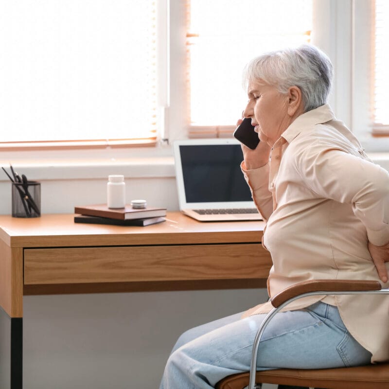 Senior woman with back pain talking by mobile phone at home